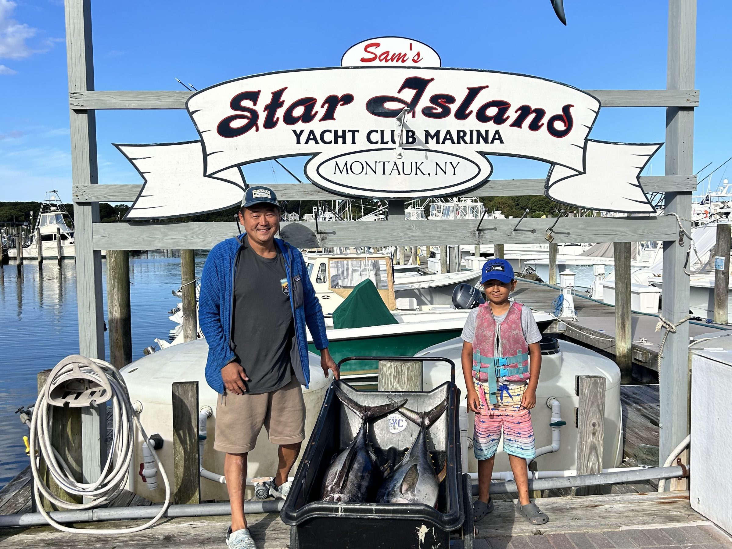 Two people with fish by dock under 'Star Island Yacht Club' sign in Montauk, NY.