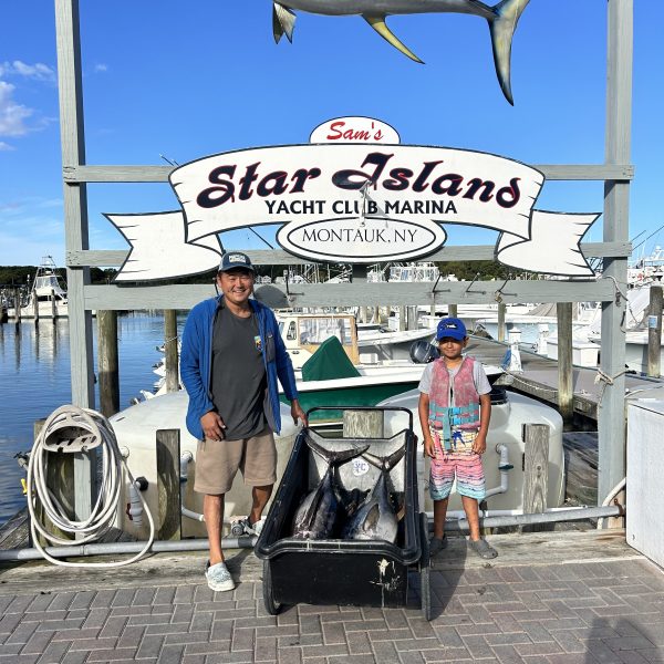 Two people with fish by dock under 'Star Island Yacht Club' sign in Montauk, NY.