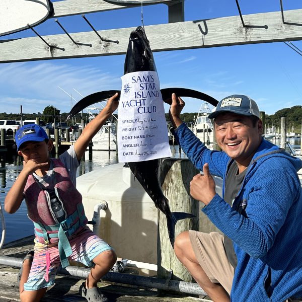 Two people posing with a fish at a dock, under a sign for Sam's Star Island Yacht Club.