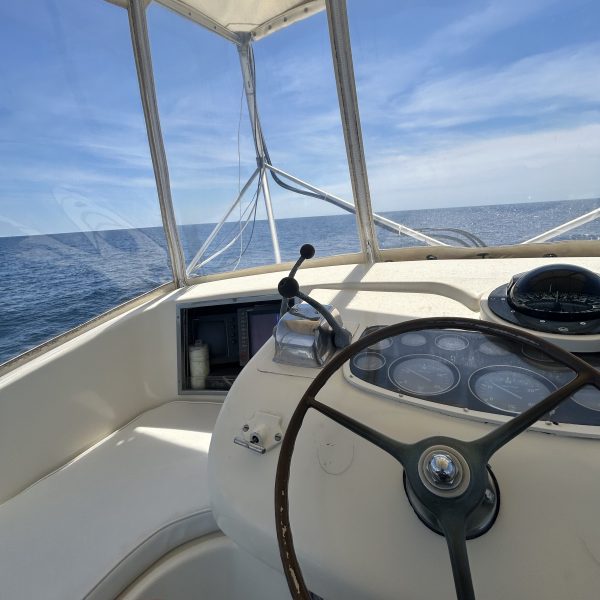 Boat cockpit with steering wheel, gauges, and ocean view under a clear blue sky.