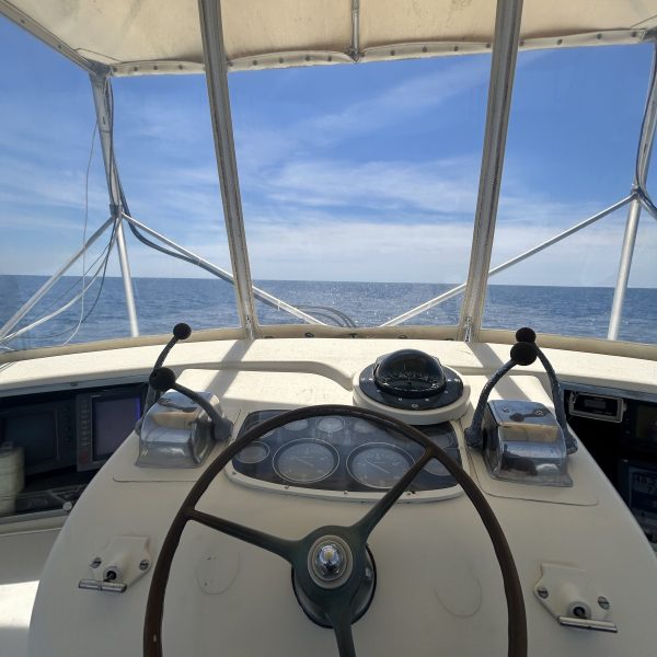 Boat helm with steering wheel and controls, ocean view in background under clear sky.