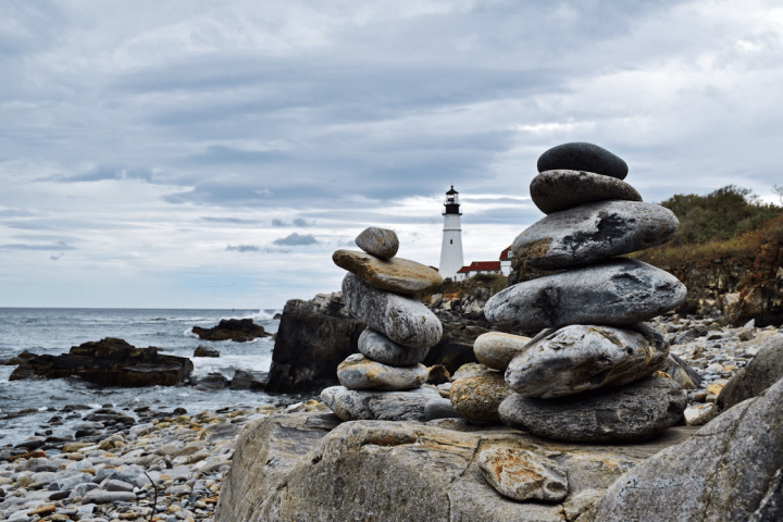 Stacked stones on rocky shore with lighthouse in the background.
