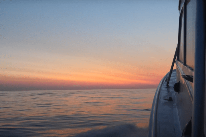 Boat sailing on calm ocean at sunset with vibrant sky colors.